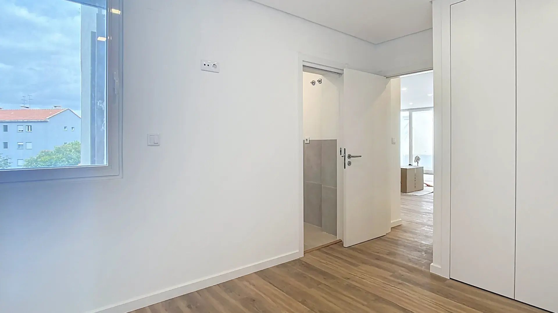 Quarto luminoso e vazio com chão de madeira, janela espaçosa e porta aberta com acesso a casa de banho privativa.