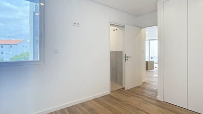 Quarto luminoso e vazio com chão de madeira, janela espaçosa e porta aberta com acesso a casa de banho privativa.