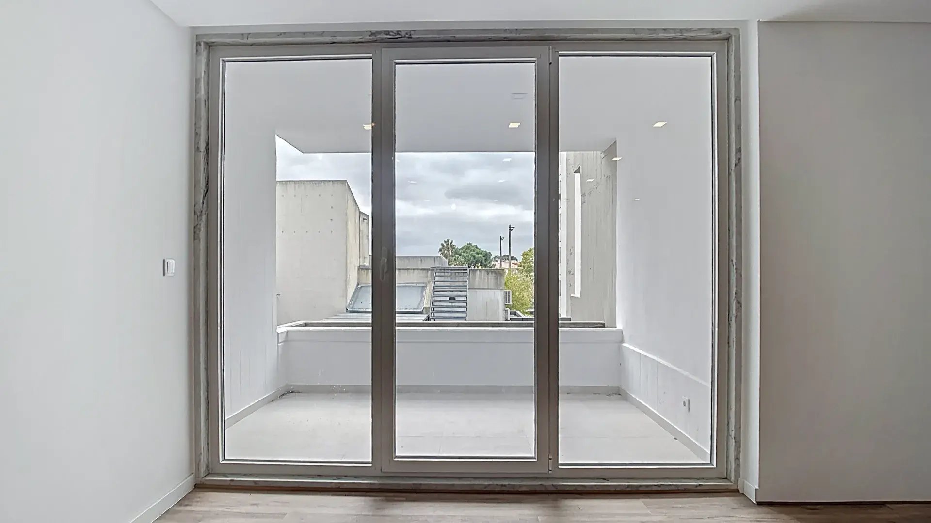 Vista interior através de grandes portas de vidro para uma varanda minimalista com paredes brancas.