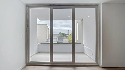 Vista interior através de grandes portas de vidro para uma varanda minimalista com paredes brancas.