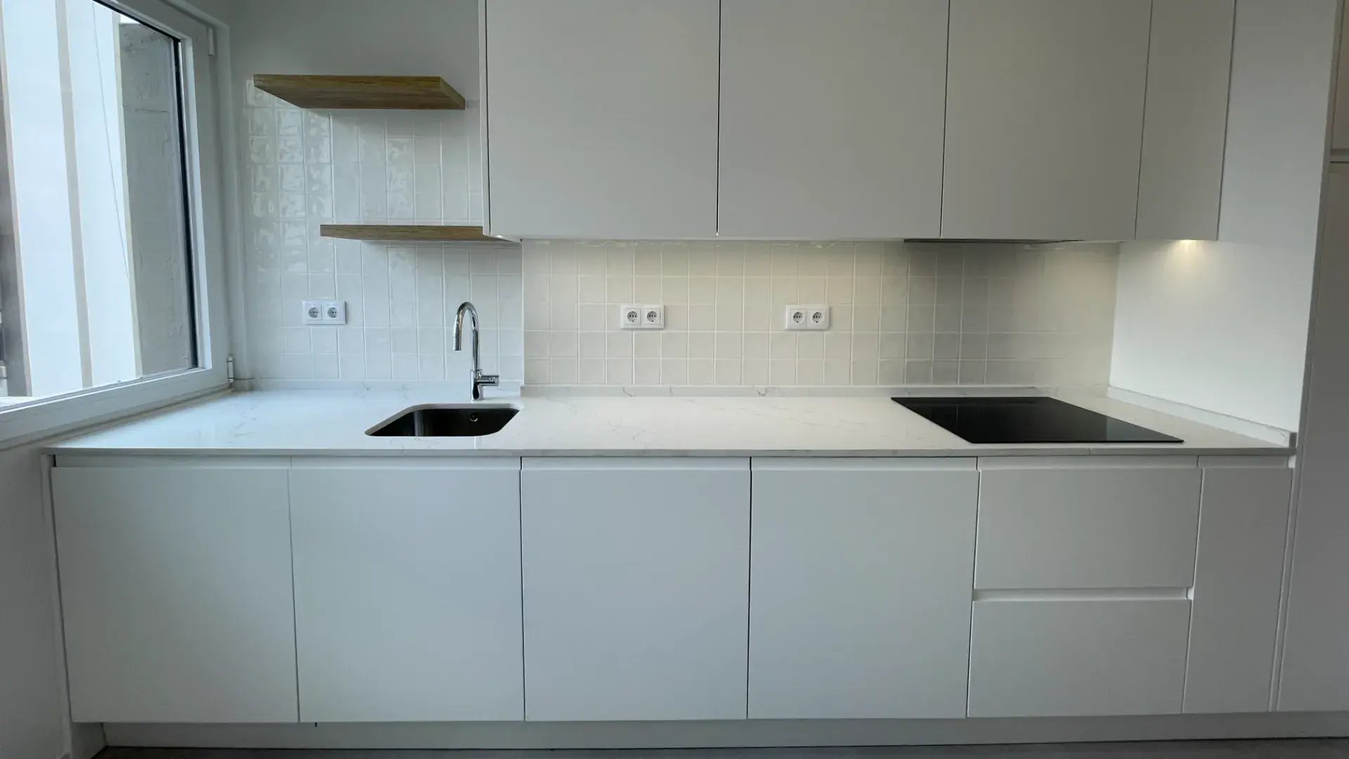Bancada de cozinha em quartzo branco com placa de indução preta, lava-louça embutido e prateleiras de madeira.